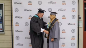A person in academic regalia adjusts the hood of a graduate in a gray cap and gown in front of a backdrop with 'Automotive Technology' and 'Rio Hondo College' logos.