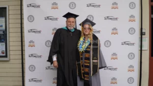 Two people in graduation gowns, one wearing a lei, stand together in front of an "Automotive Technology" step-and-repeat backdrop with "Rio Hondo College" and other logos visible.