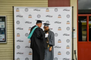 A graduate in a cap and gown receives a medal from a faculty member in regalia during a ceremony in front of a branded backdrop.
