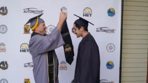 Two men in graduation caps and gowns stand in front of a backdrop adorned with logos, as one man places a stole around the other man's neck.