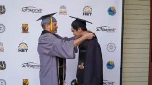 Two men in academic regalia participate in a graduation ceremony in front of a backdrop featuring logos from Rio Hondo College related to Automotive Technology and other sponsors.