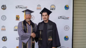Two men in graduation gowns and caps stand in front of a backdrop, shaking hands. The older man on the left smiles, while the younger man on the right has a neutral expression.