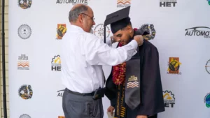 A man in a white shirt places a medal over the neck of a graduate in a black cap and gown, at a Rio Hondo College event backdrop.