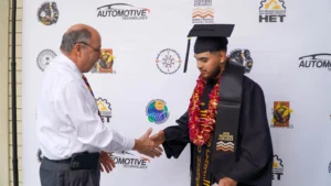 A graduate in a black cap and gown shakes hands with a man in a white shirt. The background displays logos for Automotive Technology and Rio Hondo College. The graduate wears leis and a graduation stole.