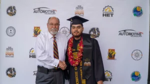 An elderly man wearing a white shirt and a young man in graduation attire pose together against a backdrop with logos of Rio Hondo College and its automotive technology program.