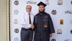 A graduate in a black cap and gown stands beside an older man in a tie. They are shaking hands in front of a backdrop with logos, including "Automotive Technology" and "Rio Hondo College.