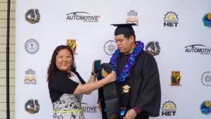 A graduate in a cap and gown receives a medal from a woman while standing in front of a banner with various logos.