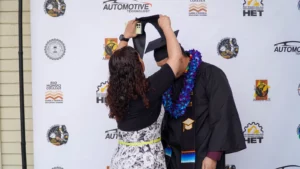 A person in a dress puts a graduation cap on another person wearing a gown and lei, standing in front of an automotive technology-themed backdrop.