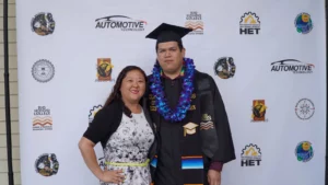 A graduate in a cap and gown with a lei around his neck poses with a woman in front of a step-and-repeat banner featuring Rio Hondo College and affiliated logos.
