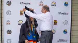 A graduate in a cap and gown receives their diploma in a ceremony. The backdrop features various logos, including "Automotive Technology" and "HET".