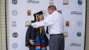 A graduate in a cap and gown stands beside a man placing a cord around her neck, in front of a backdrop with various logos and text related to an academic event.