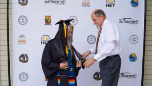 A graduate in a cap and gown stands with an older man in front of a backdrop with various logos. The man appears to be adjusting something on the graduate's gown.