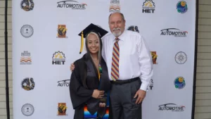 Graduate in cap and gown stands smiling next to a man with a colorful tie, against a backdrop featuring logos of institutions and organizations like Automotive Technology, Rio Hondo College, and HET.