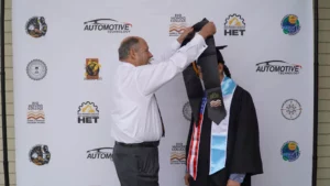 An older adult is placing a graduation stole on a younger individual dressed in a cap and gown. They are standing in front of a backdrop with multiple logos, including the Rio Hondo College logo.