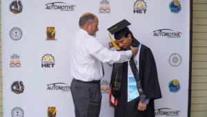 A man in a white shirt helps a graduate in a black cap and gown with a ceremonial honor cord at an event with various logos on a backdrop.