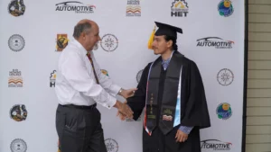 A graduate in cap and gown shakes hands with a man in a white shirt and tie in front of a backdrop with various college logos.