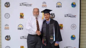 Two individuals stand in front of a backdrop featuring various logos. One is in graduation attire while the other is in business attire. They are shaking hands and smiling at the camera.