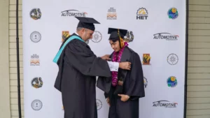 A graduate in a cap and gown receives a lei from a faculty member in front of a backdrop featuring various logos.