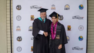 Two men in graduation gowns and caps shaking hands. One wears a lei. They stand in front of a backdrop with logos and text, including "Automotive Technology," "HET," and "Rio Hondo College.