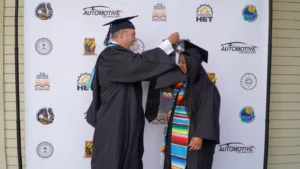 A person in graduation attire adjusts the tassel on another graduate's cap. They stand in front of a backdrop featuring various logos, including "Automotive Technology" and "Rio Hondo College.