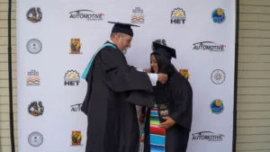 A graduate in a black cap and gown receives a certificate holder from a faculty member during a graduation ceremony, with a step-and-repeat backdrop featuring various logos behind them.