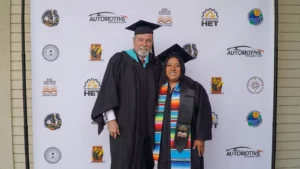 Two individuals in graduation attire stand in front of a backdrop with various logos, including "Automotive Technology." One wears a multicolored stole. Both are smiling at the camera.