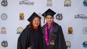 Two people wearing graduation caps and gowns pose for a photo in front of a backdrop with various educational and automotive logos.
