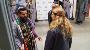 Graduates in caps and gowns converse with a woman at a ceremony, with a graduation backdrop behind them.