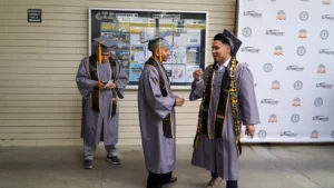 Three graduates in caps and gowns stand in front of notice boards, with two talking and one using a phone. A banner with logos is in the background.