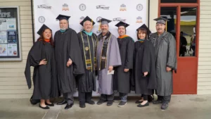 A group of seven people in graduation robes and caps stand in front of a banner on a sidewalk, smiling for a group photo.