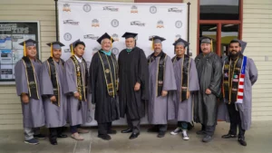 A group of people in graduation attire stands in front of a backdrop with logos. Some wear black gowns and caps, while others wear gray gowns and caps with sashes.