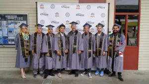 A group of eight graduates in caps and gowns stand in a line, posing for a photo in front of a backdrop with logos.