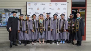 A group of graduates in caps and gowns stand in front of a backdrop with the logo "Automotive," alongside faculty members wearing academic regalia.