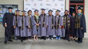 Group of graduates wearing caps and gowns standing in front of a backdrop with educators. They are posing for a photo, some holding stoles and sashes.