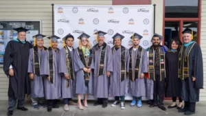 A group of graduates in caps and gowns poses for a photo in front of a backdrop with logos. Two people in suits and a person in a different gown stand with the graduates.