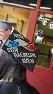 A graduation cap is decorated with miniature cars and the text "Next Stop Bachelor Ave!!" while the wearer stands near a figure in graduation attire.