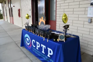 A table with a royal blue CPTP tablecloth and graduation-themed decorations is set up outside a building with double doors. The table displays gold balloons, a bowl, and informational brochures.