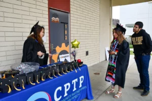 Graduates stand by a table adorned with miniature graduation caps and gifts. A woman behind the table talks to them. A large sign reads "CPTP Community Partnerships to Pipeline.