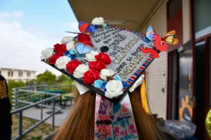 A graduate's decorated cap adorned with flowers, butterflies, and a colorful quote stands out during an outdoor event near a building and green tables.