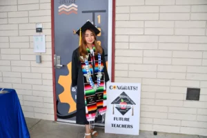 A graduate in a cap and gown stands next to a "Congrats Future Teachers" sign in front of a brick wall at a college. She wears several colorful sashes and leis.