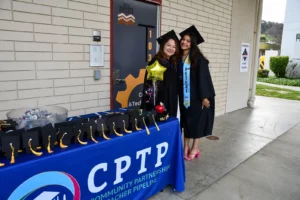 Two graduates in caps and gowns pose next to a table with miniature caps. The tablecloth reads "CPTP" for Community Partnerships Teacher Pipeline. A yellow star balloon is held by one graduate.