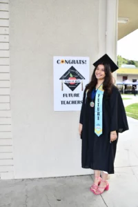 A graduate in academic regalia stands next to a sign reading "Congrats! Future Teachers". She wears a sash labeled "Future".