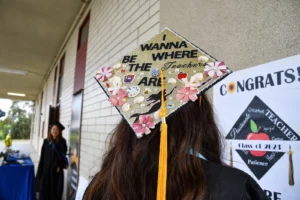 A graduate's decorated cap reads "I Wanna Be Where the Teachers Are" with various craft elements. A "Congrats" banner is visible in the background.