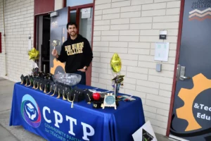 A person stands behind a booth with a blue "CPTP" tablecloth. The table features promotional items including balloons, graduation cap cutouts, and a bowl of candy. The booth is outside a building.