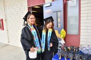 Two individuals dressed in graduation attire and "Future Teacher" sashes stand and smile near a table with decorated graduation caps.