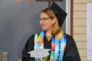 A woman wearing graduation robes and a cap stands in front of a door. She has a colorful stole with the word "TEACHER" on it. She is holding a paper cutout in the shape of a graduation cap.