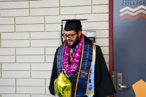 A graduate wearing a cap and gown with multiple sashes and leis stands near a door, holding a yellow balloon.