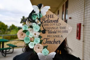 Graduation cap decorated with flowers, butterflies, and text reading "She believed she could change the world so she became a teacher.