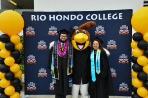 Two graduates pose with a person dressed in a bird mascot costume, standing in front of a Rio Hondo College banner and balloon columns.