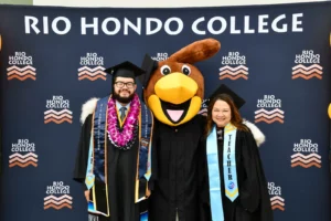 Two graduates and a person in a mascot costume stand in front of a "Rio Hondo College" backdrop. One graduate wears a lei, and the other wears a sash labeled "Teacher.